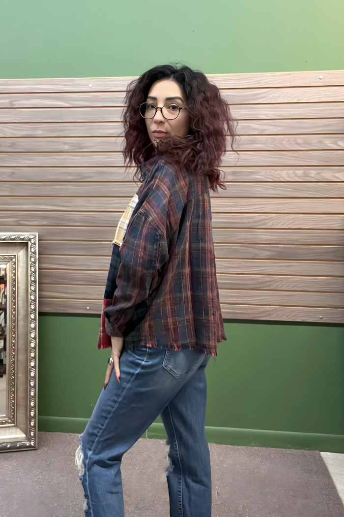 A person with wavy, shoulder-length hair and glasses stands sideways, wearing the Plaid Patchwork Button Top and blue jeans in a room with green walls, wooden panels, and a picture frame leaning nearby.
