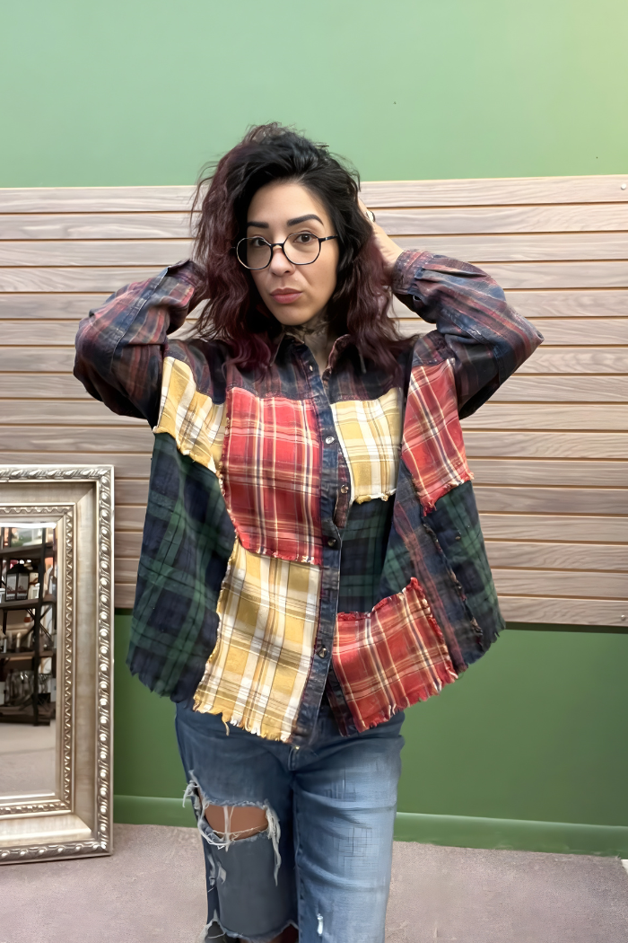 A woman with wavy dark hair and glasses poses with her hands in her hair, wearing the Plaid Patchwork Button Top and ripped jeans. She stands before a green wall with wood paneling and a framed mirror resting on the floor nearby.