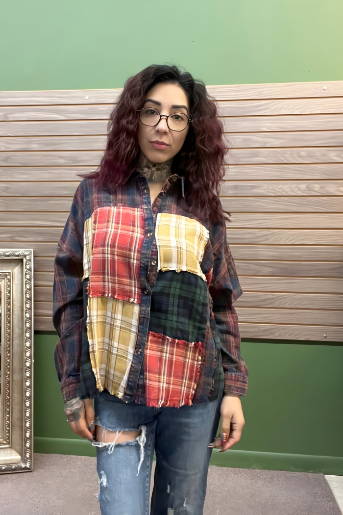 A woman with wavy, shoulder-length dark hair and glasses is indoors, wearing the Plaid Patchwork Button Top with ripped jeans. She has neck and hand tattoos, and stands against a background of wood paneling and a picture frame.