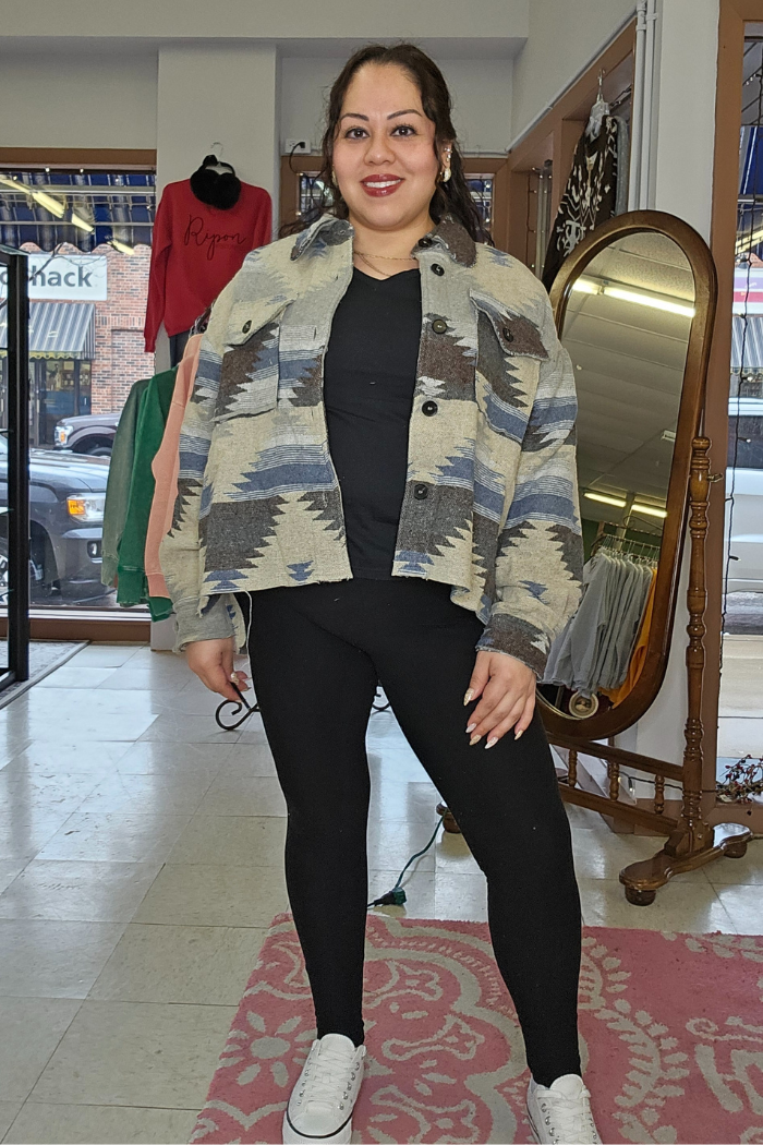 A woman stands indoors on a pink rug, smiling and wearing black leggings, a black top, white sneakers, and the Aztec Button-Down Jacket. A mirror and clothing racks are visible in the background.