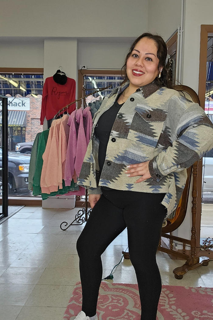 A woman wearing the Aztec Button-Down Jacket and black leggings stands smiling in a boutique, with colorful sweaters and a large mirror in the background.