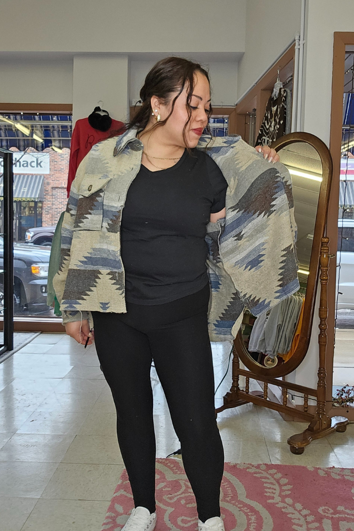 A woman in a black outfit stands in a boutique, smiling as she drapes the Aztec Button-Down Jacket over her shoulders. A mirror and clothing racks can be seen in the background.