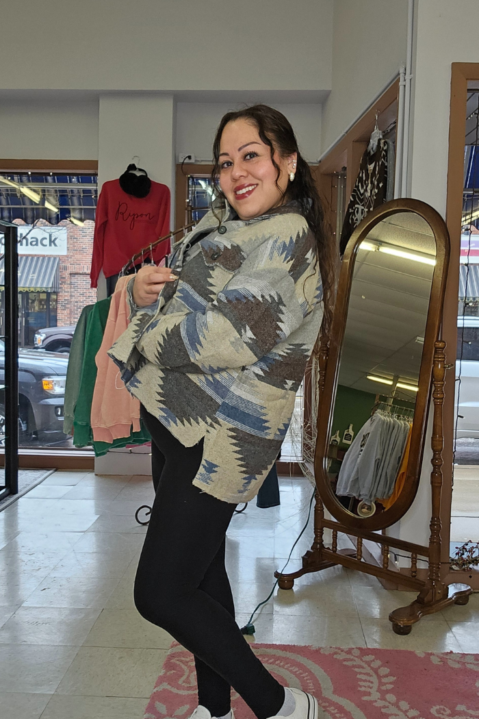 A woman smiles and poses inside a clothing store, wearing the Aztec Button-Down Jacket, black leggings, and white sneakers. A large mirror and hanging clothes are seen in the background.