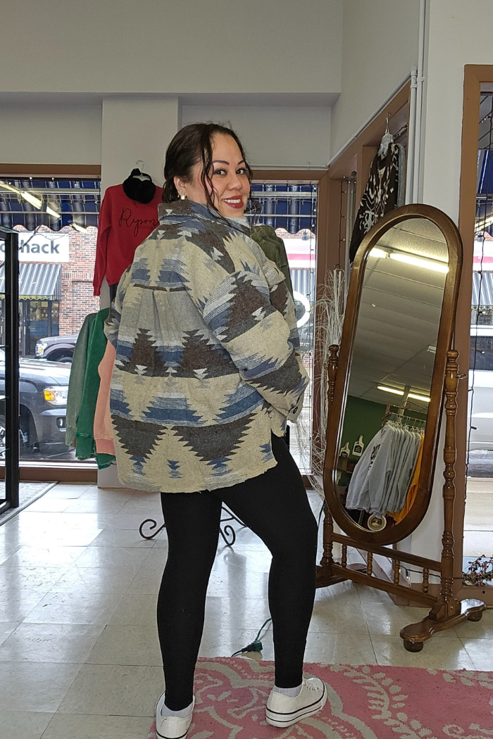 A woman stands indoors in a boutique, smiling over her shoulder while wearing the Aztec Button-Down Jacket, paired with black leggings and white shoes. A standing mirror and hanging clothes are visible nearby.