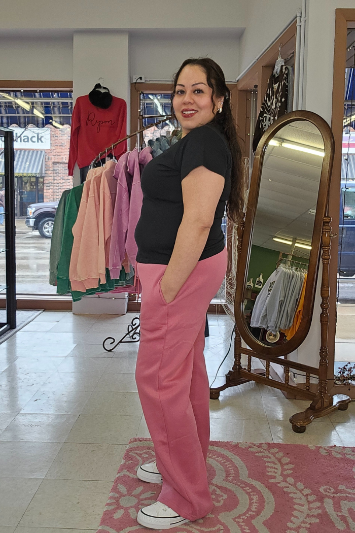 A woman with long dark hair smiles while standing sideways in a shop, wearing a black Cotton V-Neck Tee. Behind her, colorful hoodies hang on a rack near a large mirror.