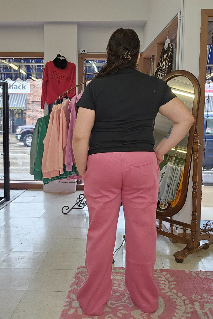 A person with long brown hair, wearing a black Cotton V-Neck Tee and pink pants, stands with their back to the camera in a clothing store. Colorful clothes hang on racks and there’s a mirror to the right.