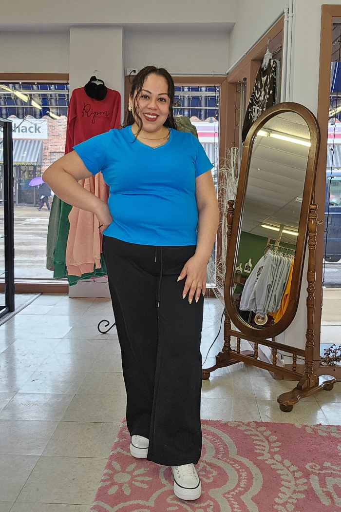 A smiling woman in a bright blue Cotton V-Neck Tee, black pants, and white sneakers stands in a boutique with clothing racks, a mirror, and a pink rug. Storefront windows reveal the street outside.