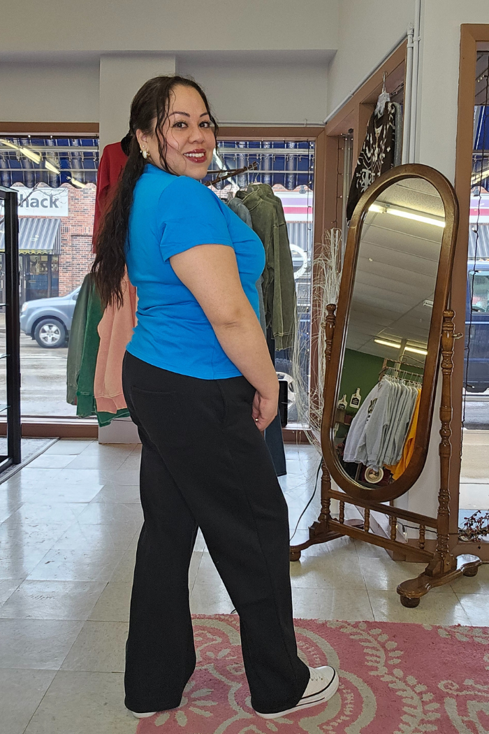 A woman with long dark hair, wearing a bright blue Cotton V-Neck Tee and black pants, smiles and poses in a clothing store near a vintage mirror and racks of clothes.