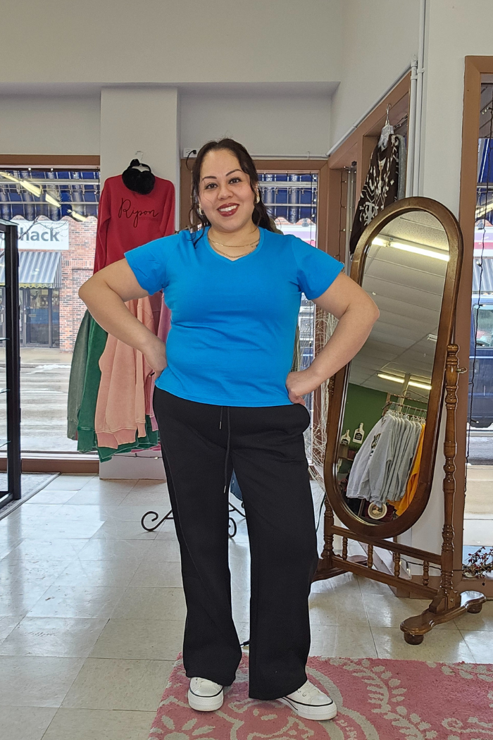 A woman smiles indoors with hands on hips, wearing a Cotton V-Neck Tee, black pants, and white shoes. Behind her are a full-length mirror, clothing rack, and large windows overlooking the street.