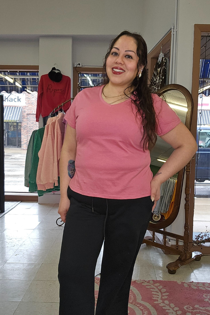 A woman with long dark hair, wearing a pink Cotton V-Neck Tee and black pants, smiles in a boutique with clothing racks, a mirror, and a large window behind her, highlighting her relaxed silhouette.