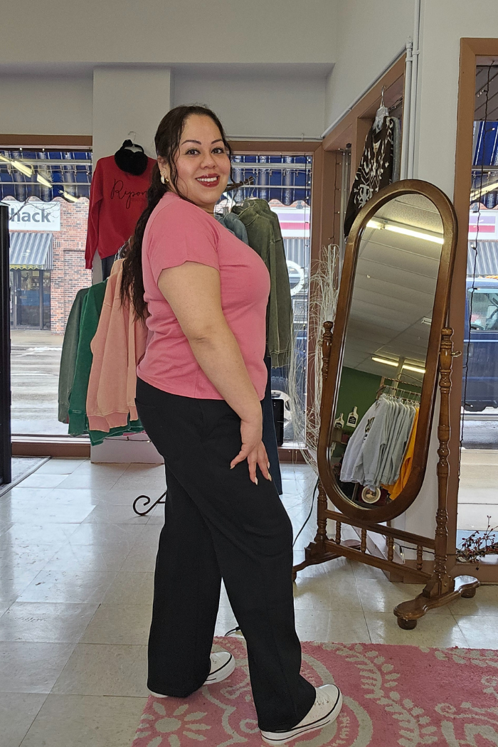 A woman with long dark hair, dressed in a Cotton V-Neck Tee, black pants, and white shoes, smiles in a boutique featuring clothing racks and a large mirror.