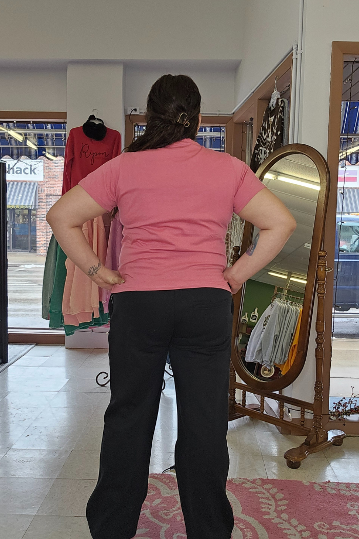 A person with long brown hair, wearing a Cotton V-Neck Tee and black pants, stands hands on hips in a boutique. Behind them are dresses, a full-length mirror, and shop windows revealing the street outside.