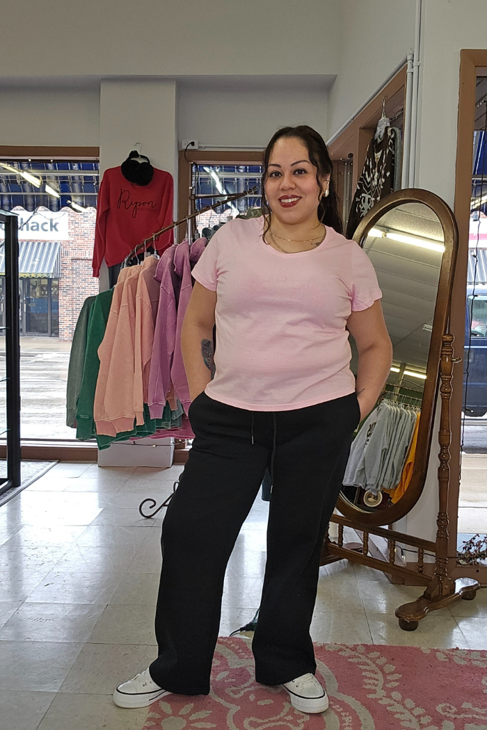 A woman smiles in a clothing store, wearing a light pink Cotton V-Neck Tee with a relaxed fit, black pants, and white sneakers. Sweatshirts hang behind her, with a standing mirror and large windows showing the street outside.