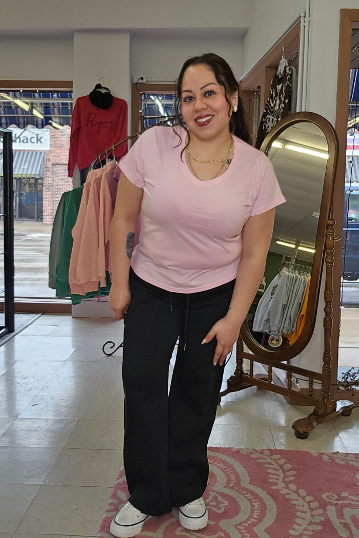 A smiling woman in a light pink Cotton V-Neck Tee, black pants, and white sneakers poses in a clothing store filled with colorful racks, a mirror, and a patterned rug. The Cotton V-Neck Tee’s relaxed fit flatters her style.