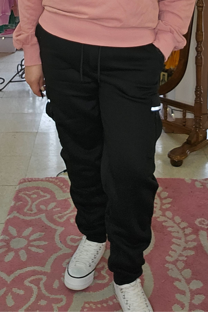 A person wearing Fleece-Lined Cargo Joggers, a pink sweatshirt, and white sneakers stands on a pink patterned rug, one hand in their pocket. Only the lower half of their body is visible.