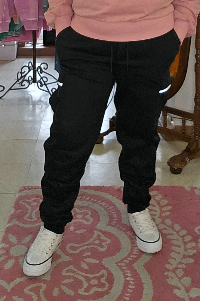 A person stands indoors on a pink patterned rug, hands in pockets, wearing Fleece-Lined Cargo Joggers, a pink hoodie, and white sneakers. Their face is not shown.