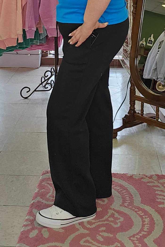 Someone wearing Wide Leg Fleece Sweatpants, a bright blue shirt, and white sneakers stands on a pink patterned rug in a clothing store, resting their hand on their hip. Clothing racks and a mirror are seen in the background.