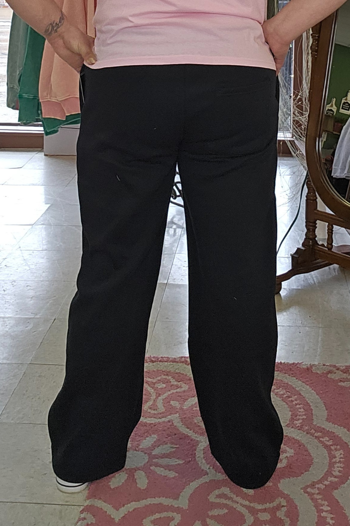 A person stands indoors with their back to the camera, wearing Wide Leg Fleece Sweatpants in black and a light pink top, hands on hips, on a patterned pink rug.