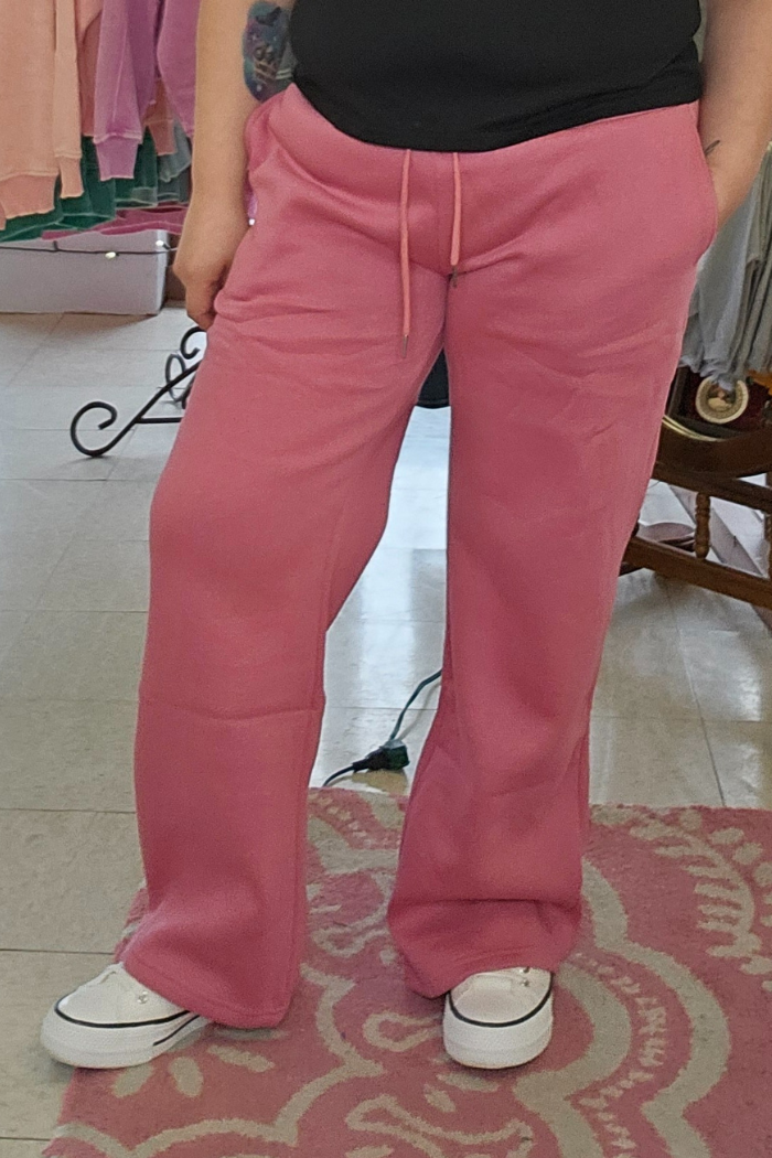 A person wearing Wide Leg Fleece Sweatpants in pink and white sneakers stands on a pink patterned rug, with only their lower half and a partial black shirt visible. Clothing and hangers are seen in the background.
