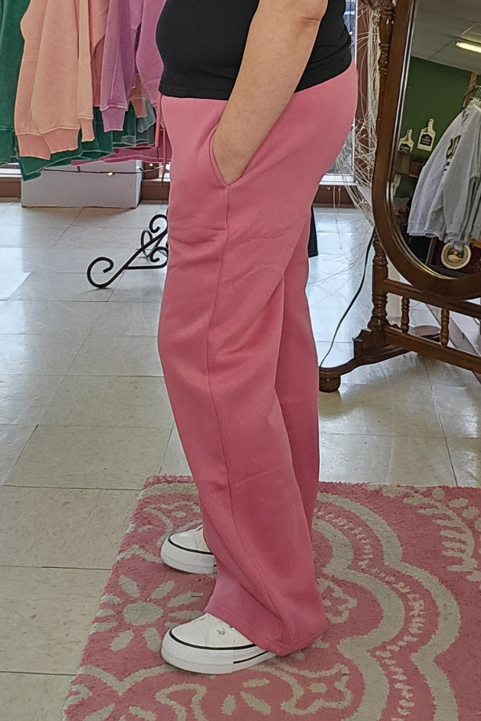 Indoors, a person wears Wide Leg Fleece Sweatpants with a black shirt and white sneakers on a pink patterned rug. Their hands are in their pockets, with more clothing visible in the background.
