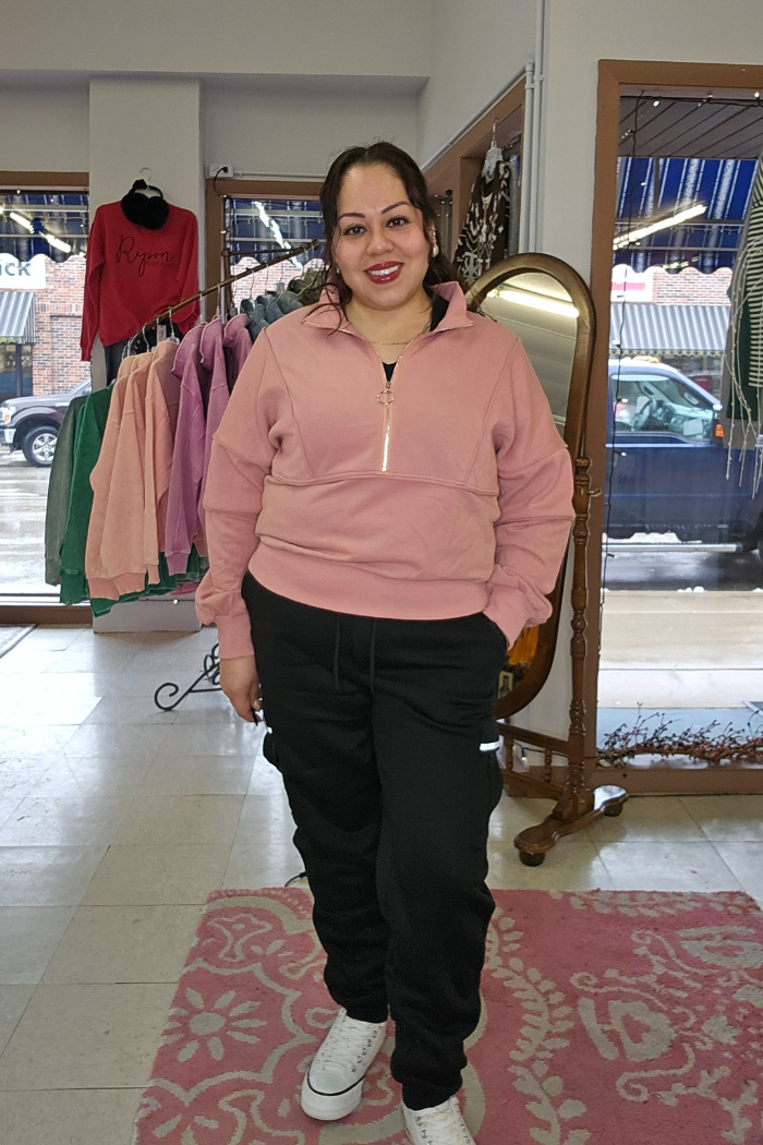 A woman smiles in a boutique, wearing a pink Half Zip Pullover Sweater, black joggers, and white sneakers. Colorful sweaters hang behind her, and a large window offers a lively street view outside.