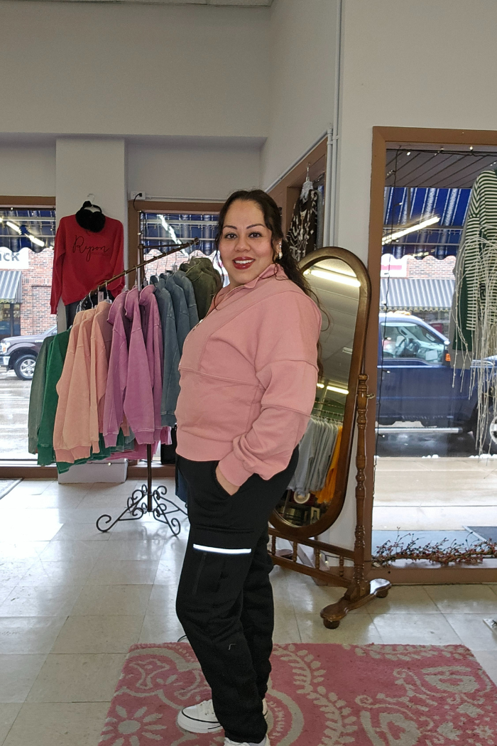 A woman wearing a dusty rose Half Zip Pullover Sweater and black pants smiles in a clothing store, with colorful sweatshirts on display and a large mirror behind her. Outside the window, cars are parked on a rainy day.