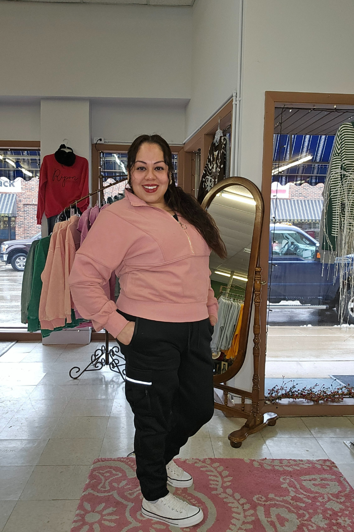 A woman with long brown hair, wearing a Half Zip Pullover Sweater in dusty rose, black pants, and white sneakers, smiles in a boutique with clothing displays, a mirror, and a window revealing parked cars outside.