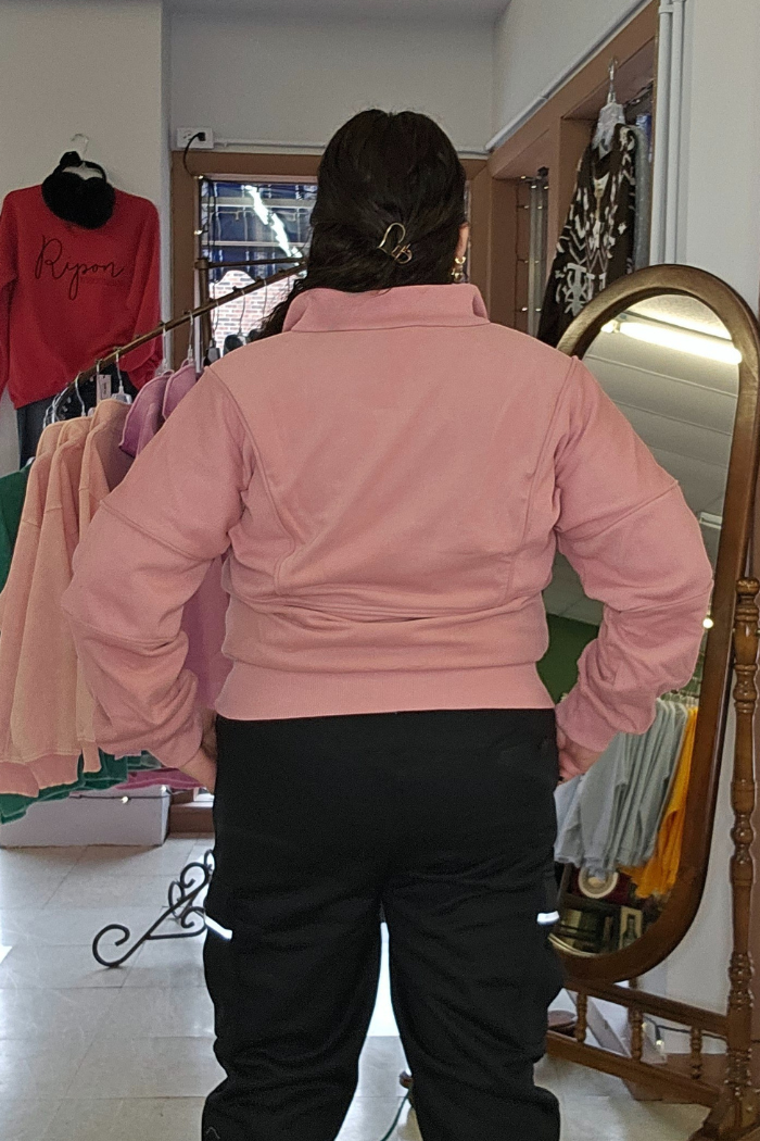 A person with dark hair in a clip stands indoors, facing away from the camera, wearing a pink Half Zip Pullover Sweater and black pants. Clothing racks and a large mirror can be seen in the background.