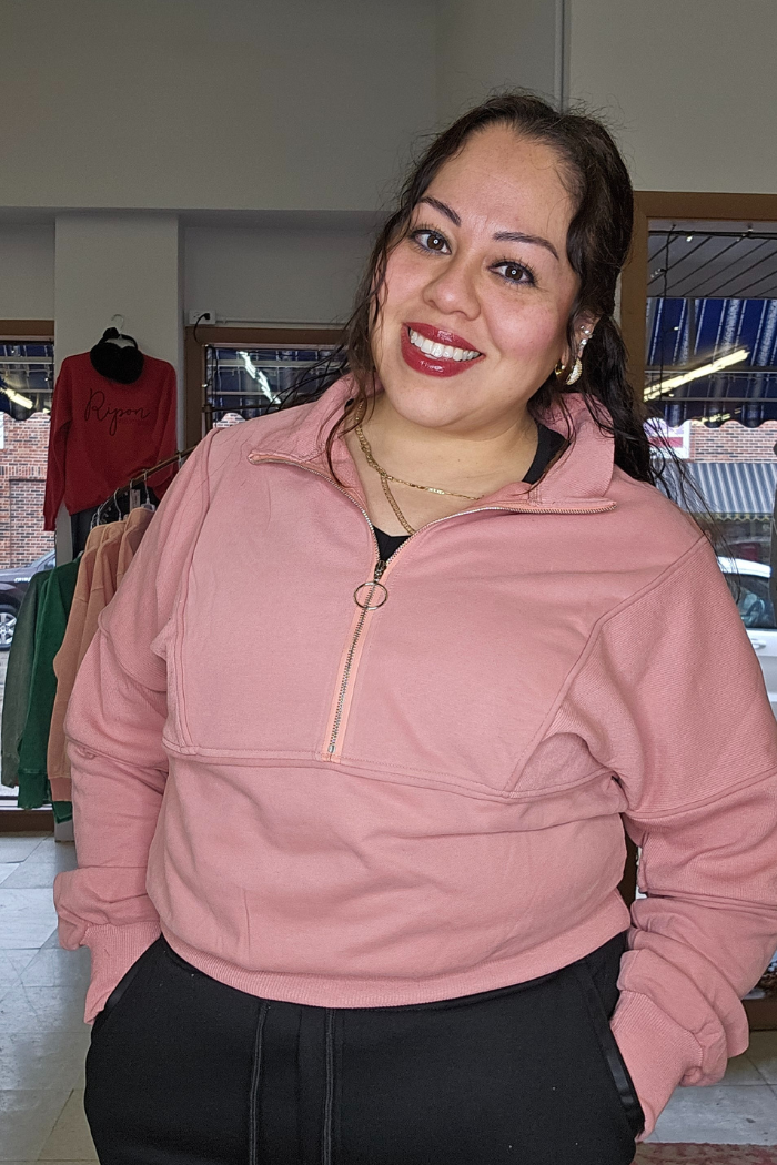 A woman with long dark hair smiles indoors, wearing the Half Zip Pullover Sweater in dusty rose and black pants. Behind her, clothing racks and a window with a street view are visible.