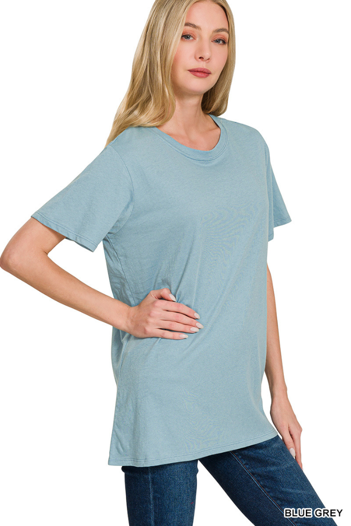 A woman with long blonde hair wears a loose-fitting Zenana Cotton Basic Tee in blue-grey and blue jeans, standing with one hand on her hip against a white background.