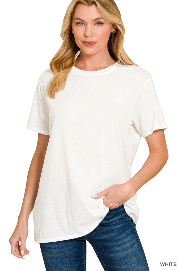 A woman with long blonde hair models the Cotton Basic Tee in a relaxed fit with blue jeans. She poses against a white background, holding the hem of her shirt with one hand and keeping the other by her side.