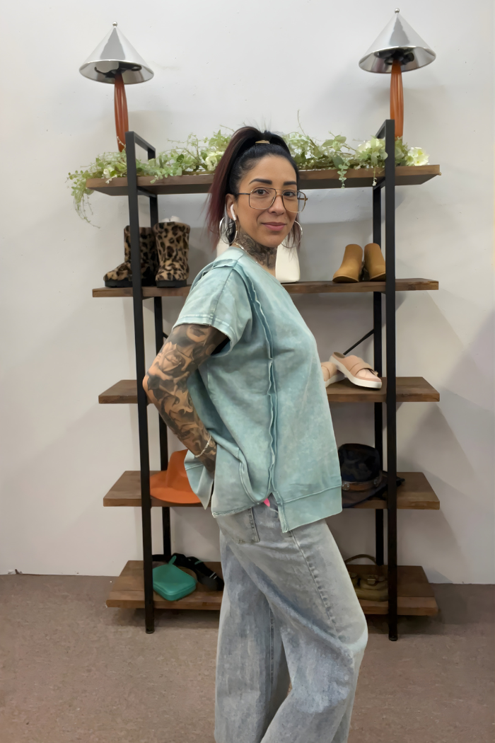 A woman with tattoos, glasses, and a ponytail smiles sideways in an Acid Wash Hi-Low Top and jeans, standing before shelves adorned with shoes, bags, plants, and two wall lamps.