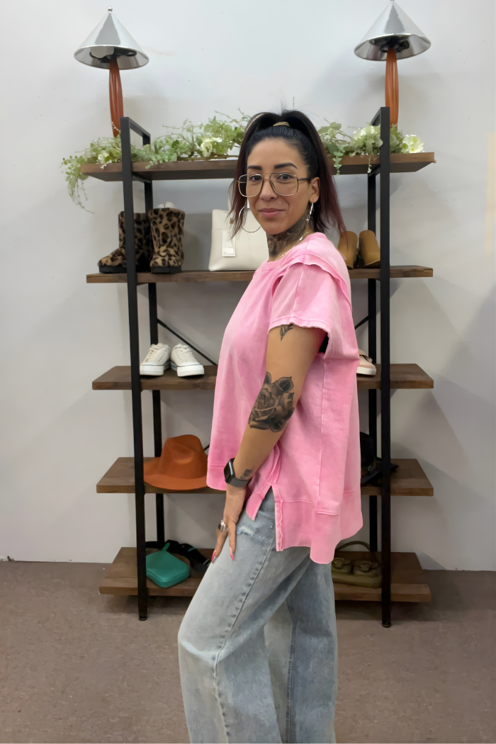 A woman with glasses and a ponytail, smiling as she stands sideways, wears a pink Acid Wash Hi-Low Top and light blue jeans. Behind her are shelves displaying shoes, plants, hats, and bags.