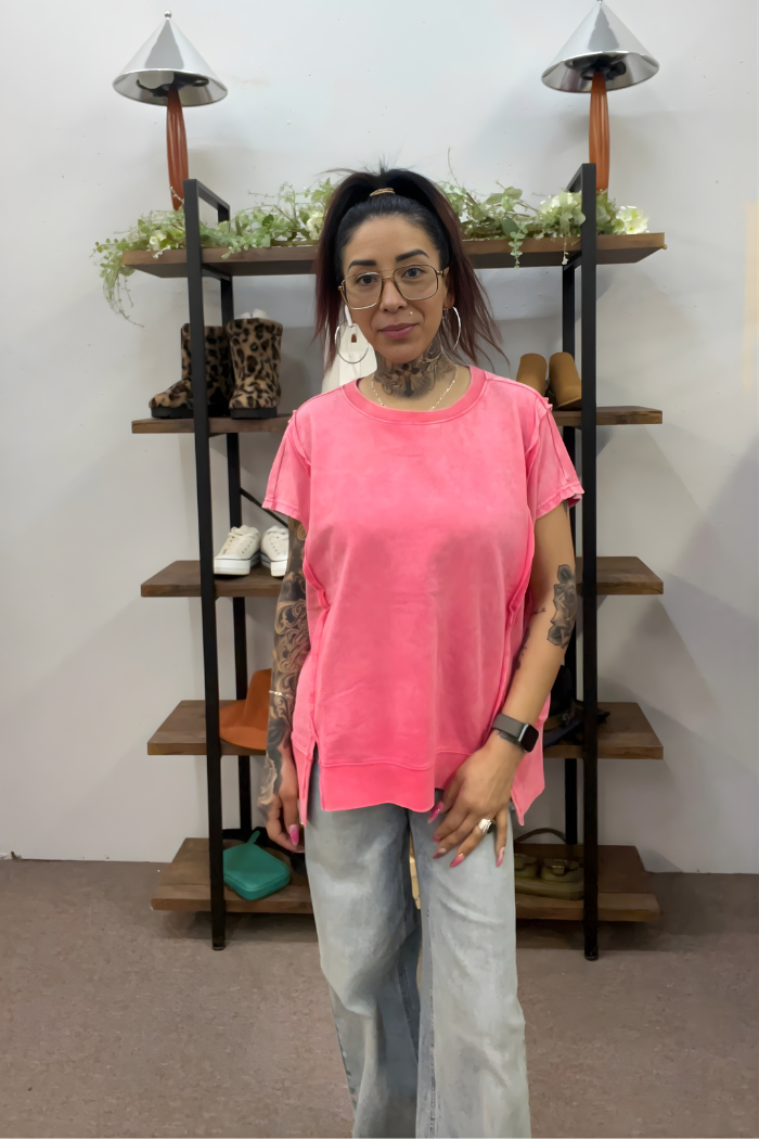 A woman with glasses and tattoos wears a bright pink Zenana Acid Wash Hi-Low Top and light blue jeans, standing indoors in front of a black shelving unit displaying shoes, purses, and greenery.