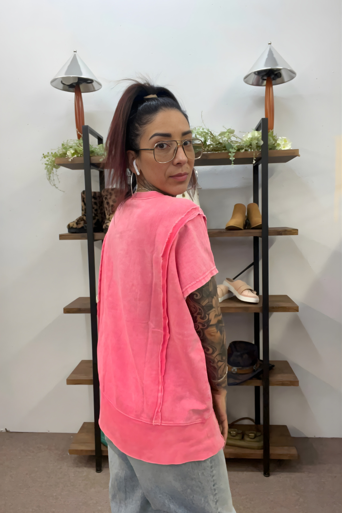 A woman with glasses and a high ponytail, wearing a pink Acid Wash Hi-Low Top and light jeans, stands by a shelving unit with shoes and decor, looking over her shoulder at the camera.
