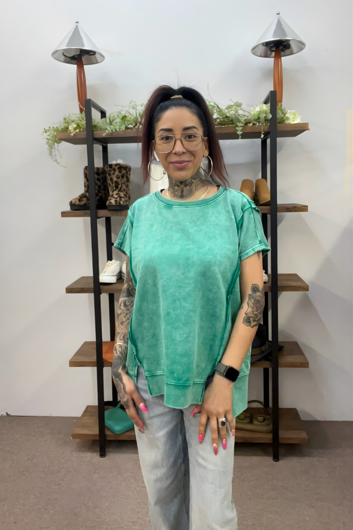 A tattooed woman with glasses and a ponytail wears a green Acid Wash Hi-Low Top and light jeans, smiling in front of a shelving unit displaying shoes, plants, and decor.