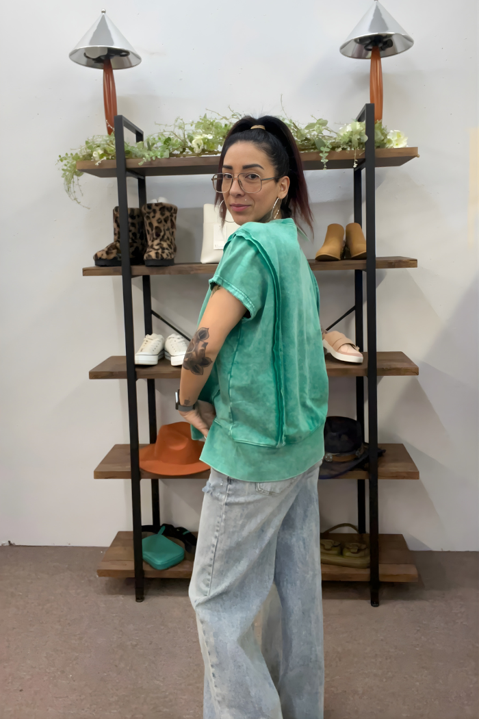 A woman in an Acid Wash Hi-Low Top and light blue jeans stands before a shelf with shoes, hats, and plants. She glances over her shoulder at the camera, showcasing a tattoo on her arm, a ponytail, and effortless style.