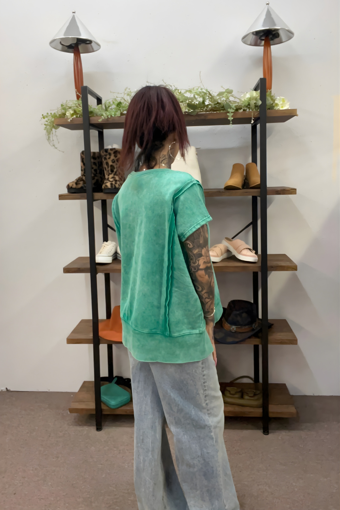 A tattooed individual wearing an Acid Wash Hi-Low Top and light blue jeans stands with their back to the camera in front of a shelving unit displaying shoes, bags, and hats.