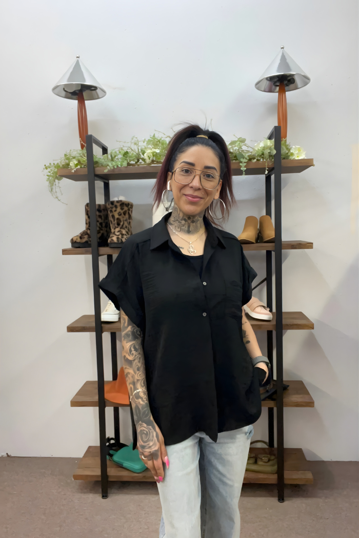 A woman with glasses and tattoos, wearing the Airflow Button Down in black and light jeans, smiles while standing before shelves filled with shoes, bags, and decorative greenery.
