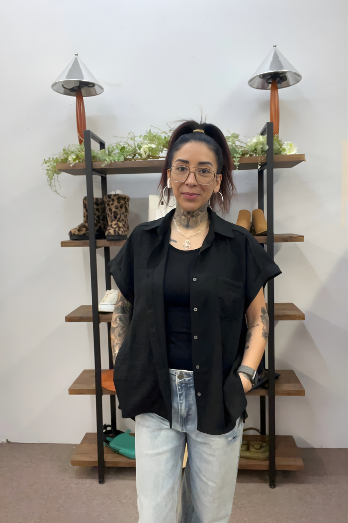 A tattooed woman with glasses and a ponytail stands indoors by shelves with shoes and plants, wearing the lightweight Airflow Button Down over a black top and light jeans for an easy, casual style.