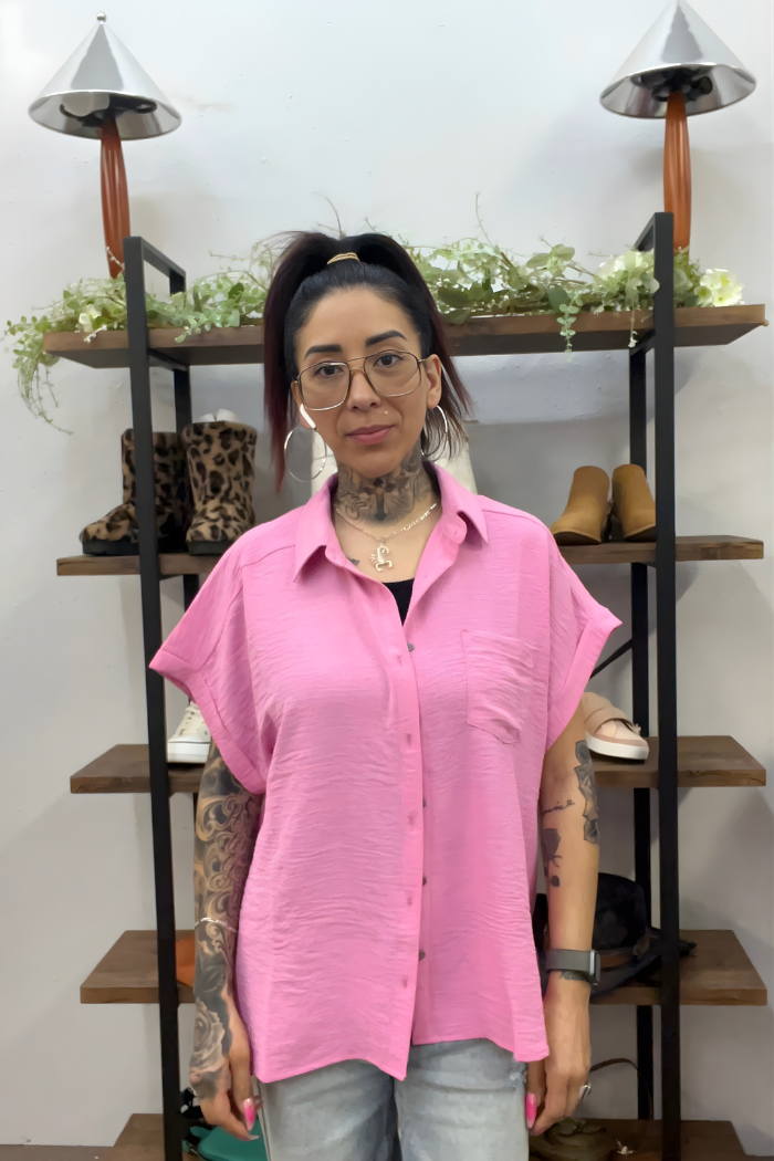 A woman with tattoos and glasses wears the pink Airflow Button Down and light jeans, standing by a wooden shelf with shoes and plants—a perfect showcase of this casual wardrobe staple.