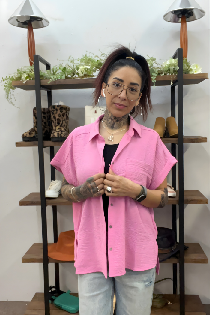 A woman with tattoos and glasses, in a pink Airflow Button Down shirt and light jeans, stands by shelves with shoes and plants. Her hair is in a ponytail, and she wears earphones and jewelry.