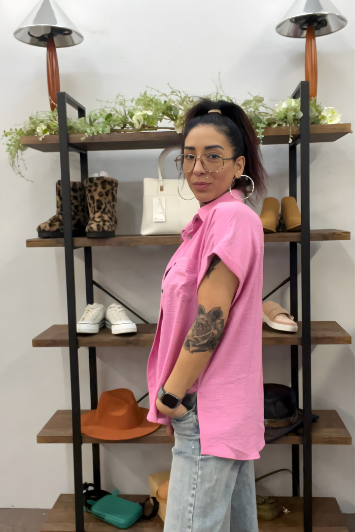 A woman with glasses and a ponytail wears the Airflow Button Down in pink with jeans. She stands sideways by a wooden shelf of shoes, bags, and hats, showing a large forearm tattoo as she looks at the camera.