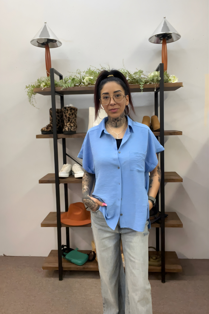 A woman with tattoos, glasses, and a ponytail is indoors wearing the Airflow Button Down in blue with light jeans. Behind her, a wooden shelf holds shoes, hats, bags, and small plants.