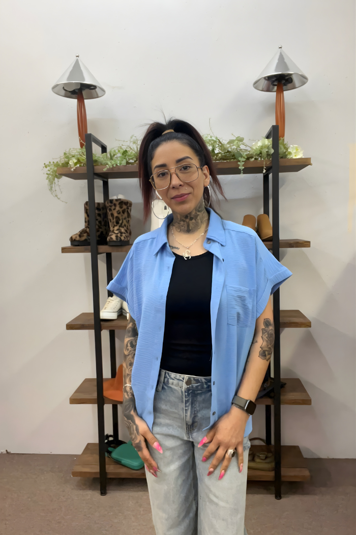 A woman with long dark hair, glasses, and tattoos stands indoors wearing the Airflow Button Down over a black top and jeans. Behind her, a wooden shelf displays shoes, plants, and lamps for a stylish backdrop to her casual look.
