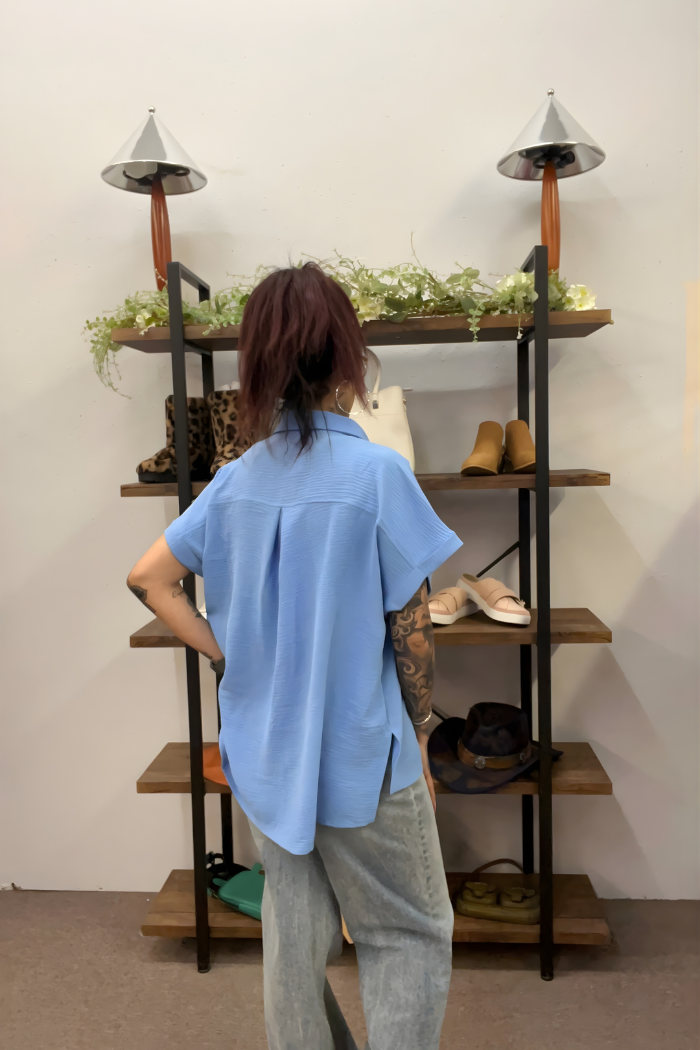 Wearing the Airflow Button Down and faded jeans, a person with long, dark hair in a ponytail stands facing shelves displaying shoes, bags, hats, and decorative greenery.