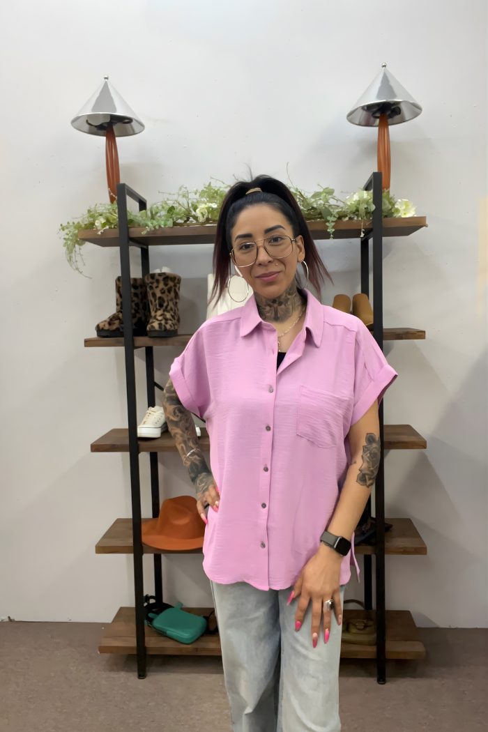 A woman with tattoos, glasses, and a ponytail stands indoors wearing the pink Airflow Button Down and light blue jeans. Behind her are shelves displaying shoes, hats, greenery, and decorative lamps.