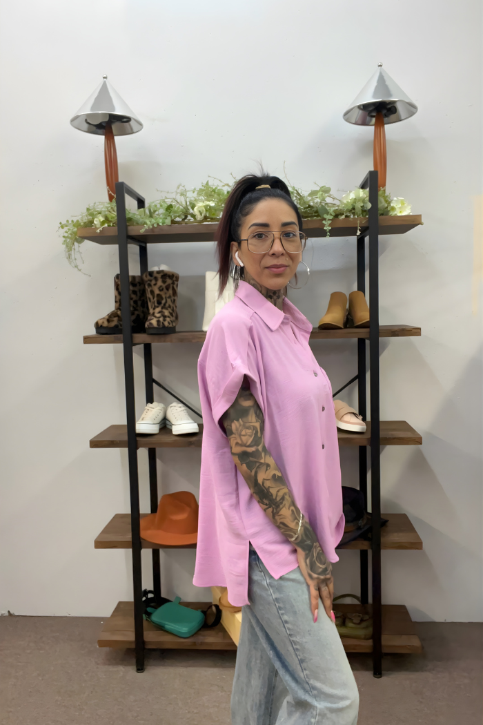 A woman with glasses and tattoos wears a lightweight pink Airflow Button Down and light jeans, standing sideways by shelves of shoes, hats, and greenery. She looks at the camera with a slight smile.
