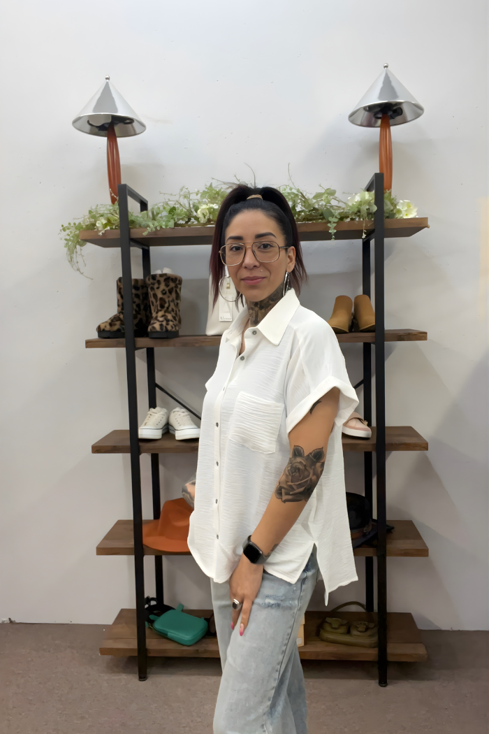 A woman with glasses, a ponytail, and a forearm tattoo smiles slightly while wearing the Airflow Button Down and light jeans, standing before a wooden shelf displaying shoes, hats, and small bags.
