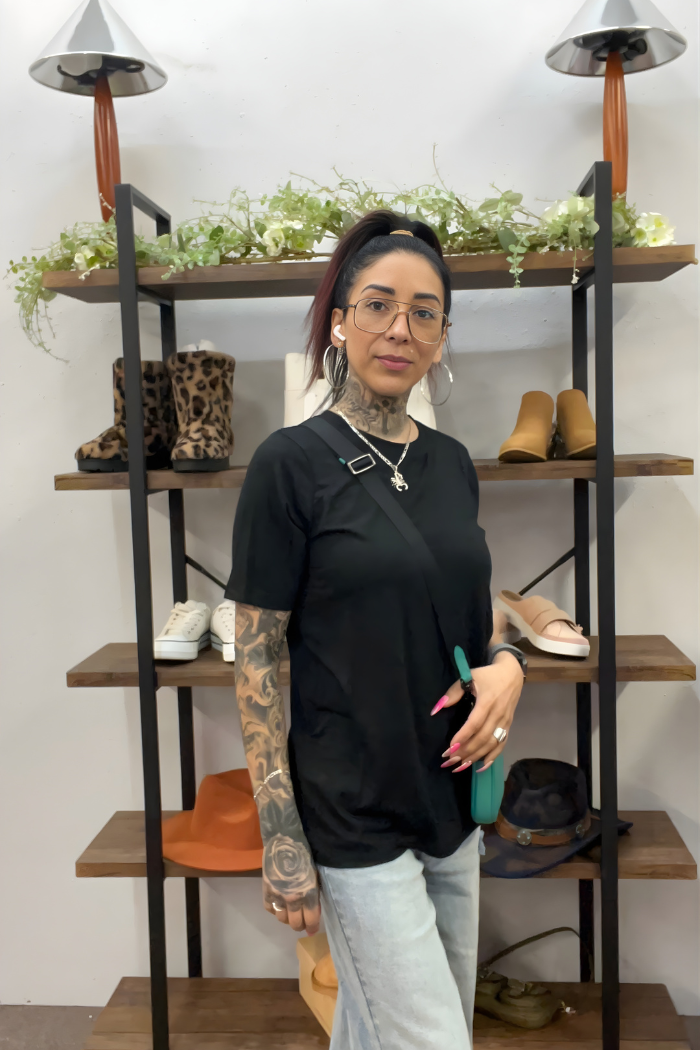 A woman with glasses and tattoos stands in front of shelves with shoes, hats, and plants. She wears the Zenana Basic Short Sleeve Tee, light jeans, and has a small crossbody bag.